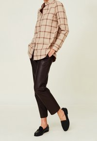 Camisa de cuadros beige con detalles marrones, combinada con pantalones de cuero negros ajustados y mocasines negros, con un diseño acortado en los tobillos.
