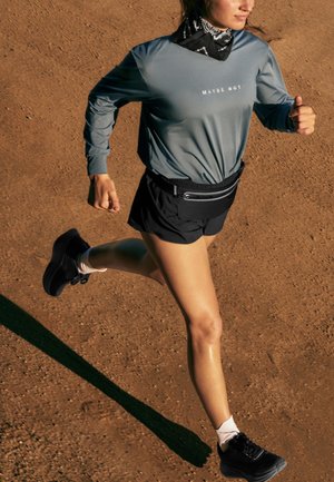 Tekač nosi črne kratke hlače, sivo dolgo majico z napisom "MAYBE NOT", črne tekaške copate in črno pasno torbico na zemeljski poti.