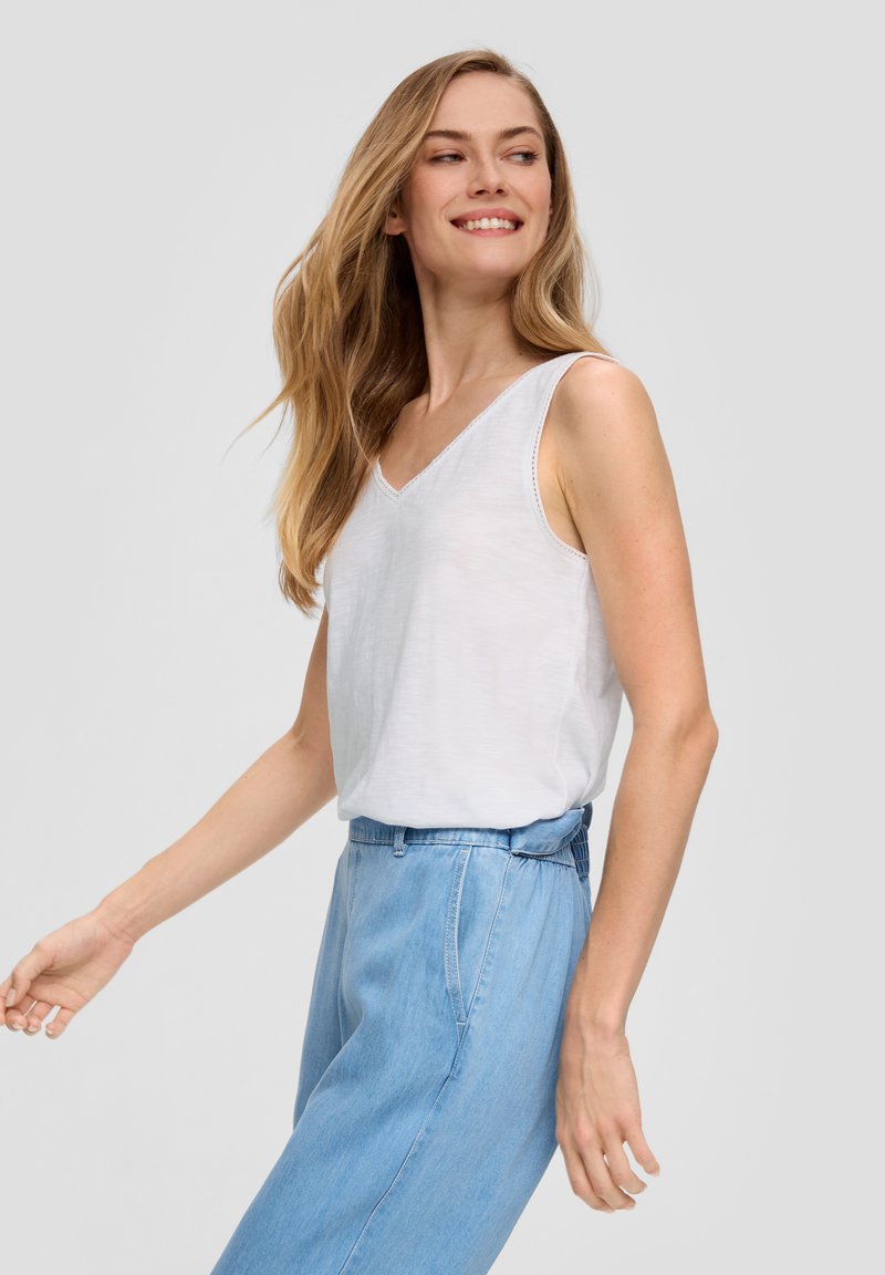 Witte mouwloze top met een V-hals en subtiele textuur, gecombineerd met lichtblauwe high-waisted jeans. Eenvoudig en modern ontwerp.