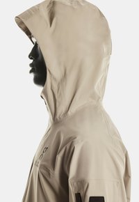 Lichtbeige waterdichte jas met capuchon, gladde textuur, zijzak met rits, logodetail op de mouw en een getailleerde pasvorm.