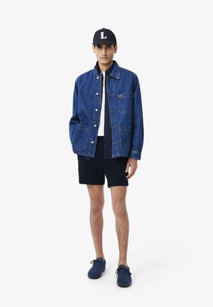 Giacca di jeans blu con due tasche frontali, abbinata a una camicia bianca, pantaloncini blu navy e sneaker blu abbinate. Il cappello presenta un logo bianco.