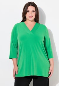 Top verde a scollo a V realizzato in tessuto morbido con maniche a 3/4, caratterizzato da una vestibilità rilassata e una texture liscia. Abbinato a pantaloni neri.