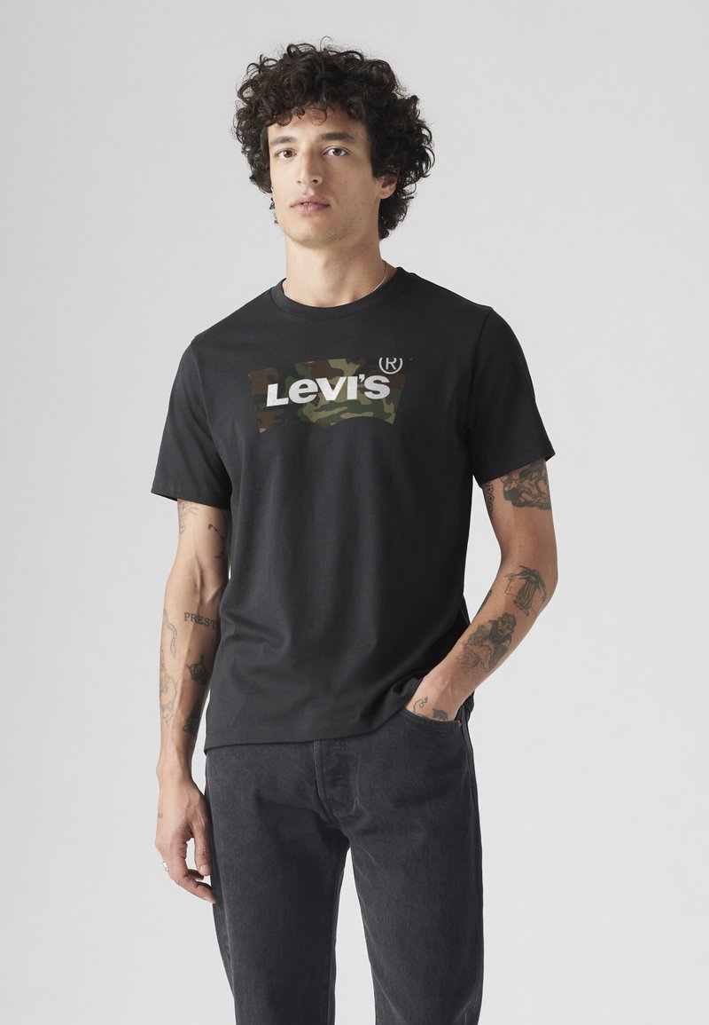 Άνδρας με σγουρά μαλλιά που φοράει μαύρο t-shirt Levi's με λογότυπο καμουφλάζ και μαύρα τζιν, στέκεται με το ένα χέρι στην τσέπη, μπροστά σε απλό φόντο.