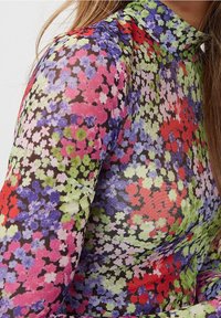 Blommig långärmad topp i genomskinligt material med ett flerfärgat mönster av röda, rosa, gröna och blå blommor mot en svart bakgrund.