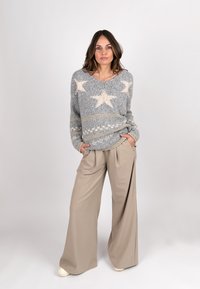 Grauer Strickpullover mit beigem Sternenmuster, V-Ausschnitt und gerippten Bündchen, kombiniert mit beigen weiten Hosen und hellen Schuhen.