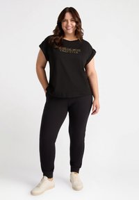 Schwarzes T-Shirt mit kurzen Ärmeln und einem goldenen Druckschriftzug auf der Vorderseite, kombiniert mit schwarzen Jogginghosen und hellen Sneakers.
