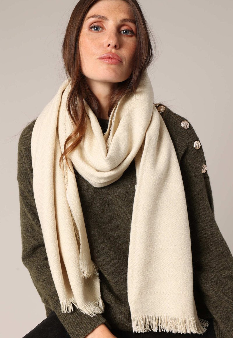 Écharpe crème au toucher doux, arborant un motif en chevrons et des bords frangés, portée sur un pull sombre.