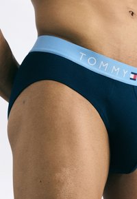 Námornícké modré slipy vyrobené z mäkkej tkaniny s svetlomodrým pásom, na ktorom je text a logo "TOMMY". Detailný pohľad zvýrazňuje textúru a strih.