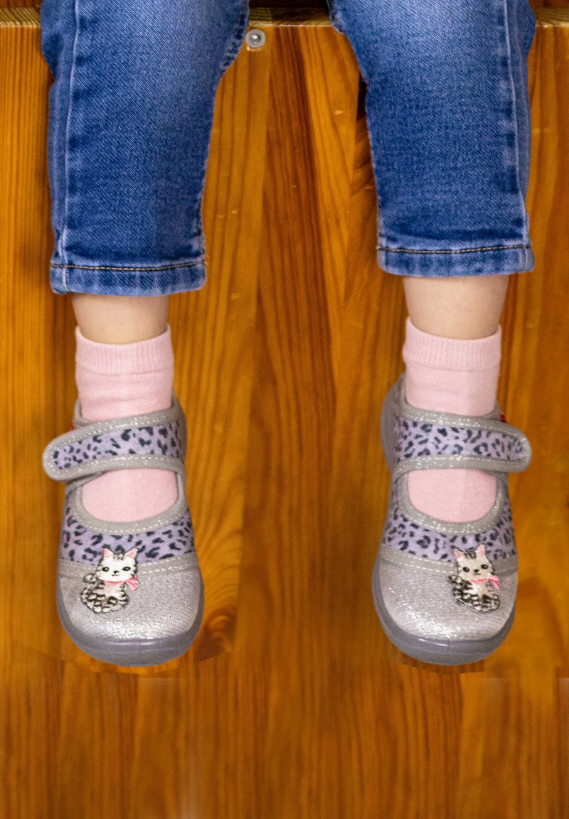 Child wearing pink socks and purple cat-themed shoes with Velcro straps, sitting with legs dangling over wooden floor.