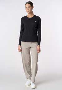 Maglietta nera a maniche lunghe con scollatura rotonda e logo dorato, abbinata a pantaloni beige a vestibilità ampia e sneakers bianche.