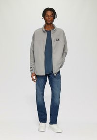 Grijze fleece jas met een hoge kraag en rits, gecombineerd met een blauwe t-shirt en blauwe spijkerbroek. Witte sneakers maken de outfit af.