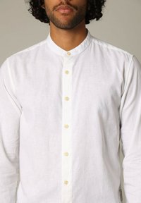 Witte linnen blouse met een bandkraag, sluiting met knopen aan de voorkant met beige knopen, lange mouwen en een subtiele textuur.