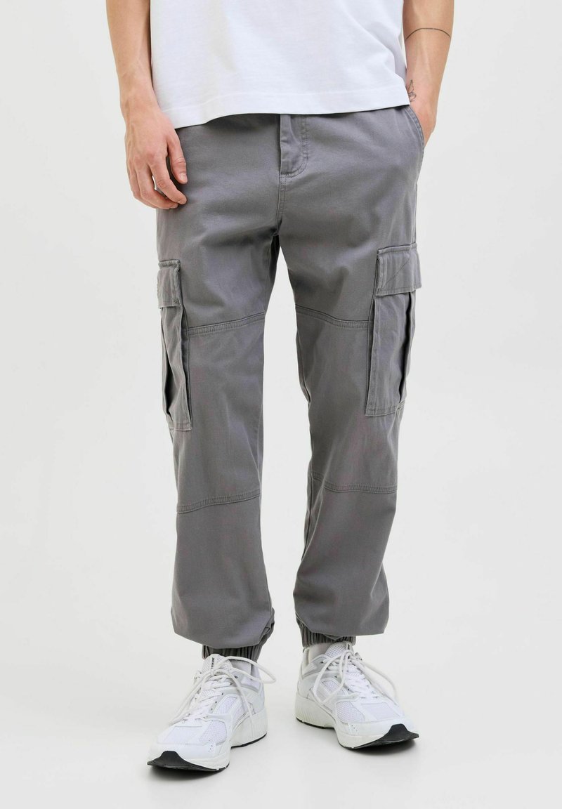 Graue Cargo-Hosen mit elastischen Bündchen, mehreren Seitentaschen und einem entspannten Schnitt, kombiniert mit weißen Sportschuhen. Baumwollmaterial.