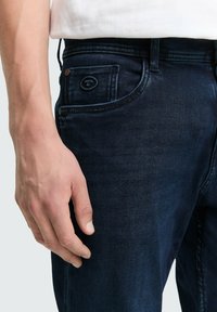 Tmavomodré denimové nohavice s klasickým strihom. Funkcie zahŕňajú malú zadnú vrecko s kruhovým logom a čistú úpravu. Ruka spočíva na stehne.