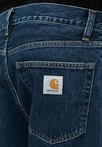 Οπίσθια τσέπη σκούρου μπλε τζιν με ορατό καφέ ράψιμο και ένα λευκό ταμπελάκι με κίτρινο λογότυπο Carhartt.