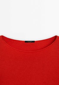Pull en maille rouge avec un col rond, présentant un tissu texturé et une étiquette noire indiquant le nom de la marque.
