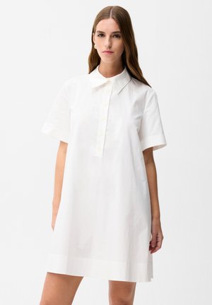 Jonge vrouw met lang bruin haar die een loszittende witte korte mouwen overhemdjurk draagt met een kraag en knoopsluiting, tegen een witte achtergrond.