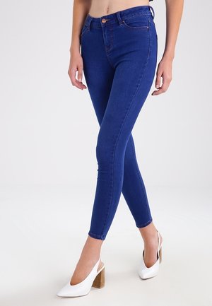 Vrouw met een blauwe skinny jeans en witte hoge hakken met slingback die met gekruiste benen staat.