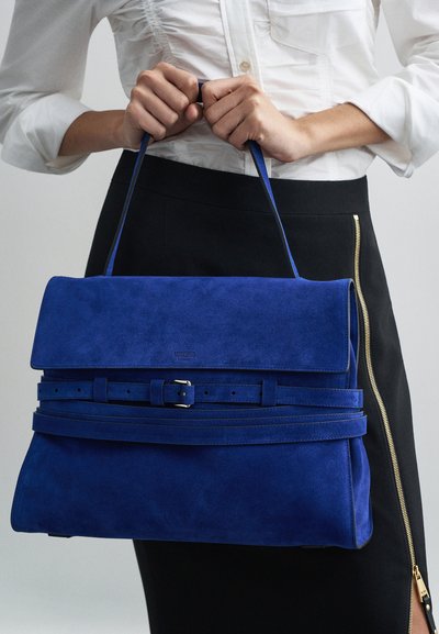 Τσαντάκι ώμου από μπλε σουέντ με δομημένο σχήμα, επίπεδη καπάκι, ζωνάκι και λεπτομέρεια λογότυπου. Ο ιμάντας είναι ρυθμιζόμενος, συμπληρώνοντας ένα κομψό σύνολο.