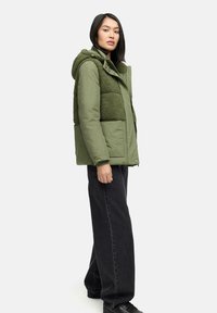 Groen gewatteerd jack met een textuur, fleece-achtig voorpaneel en capuchon, met drukknopen. Gekozen in combinatie met zwarte wijde jeans.