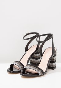 Musta patentoitu nahka korkeakorkoiset sandaalit, joissa on koristeellinen strassihihna ja lohkokorko, joka esittelee raidallista metallikuviota.