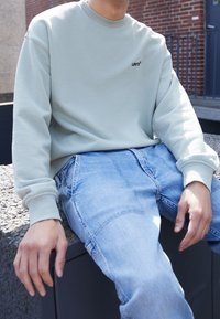 Joven sentado al aire libre con sudadera Levi's verde claro y jeans de mezclilla azul claro, frente a una pared de ladrillos.