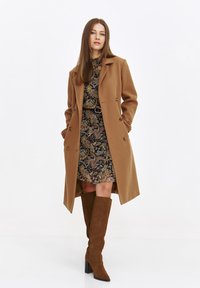 Brauner Wollmantel mit doppelt geknöpften Knöpfen, über einem gemusterten Kleid und braunen kniehohen Wildlederstiefeln mit Blockabsätzen, stehend vor einem weißen Hintergrund.