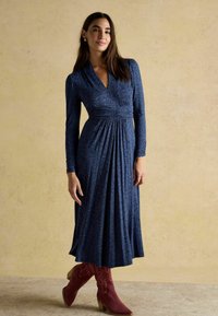 Tumesinine pikade käistega kleit V-kaelusega, kogutud vöökoha ja voogava seeliku ning burgundia kuldnokka saapadega, halli tekstuuriga taustal.