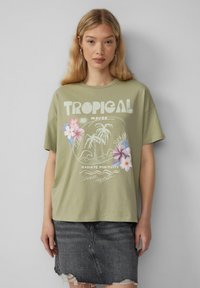 T-shirt en coton vert olive à manches courtes, avec un design graphique tropical comprenant des fleurs et des palmiers, associé à une jupe en denim noire.
