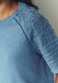 Blauw shirt met korte mouwen met getextureerde kantdetails op één schouder. Zachte stof met een licht gemêleerd uiterlijk.