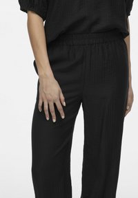 Melni, teksturēti auduma bikses ar elastīgu jostu, ar brīvu piegriezumu un taisnu kāju dizainu. Roku atpūta uz bikses kājas.