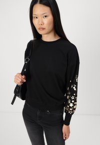 Svart sweatshirt med lange, oversized ermer som har gullfargede blomstermønstre. Ribbestrikket mansjett og nederkant, løs passform, laget av mykt stoff.