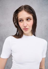 Weißes, kurzärmliges, figurbetontes Oberteil mit Rundhalsausschnitt. Glatte Textur mit nahtlosem Design. Das Model hat nasses, welliges Haar und ist leicht zur Seite positioniert.