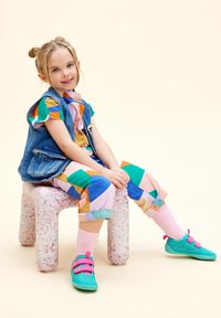 Conjunto colorido con un chaleco de mezclilla, pantalones estampados en rosa, verde y azul, combinados con zapatos color aqua y calcetines rosas, sentado en un taburete texturizado.