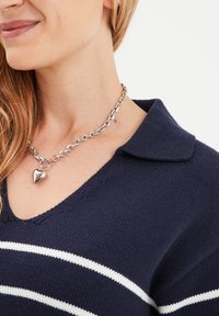 Navy gebreide trui met witte strepen, V-hals en een gestructureerde kraag. Heeft een zilveren kettinghalsketting met een hartvormige hanger.