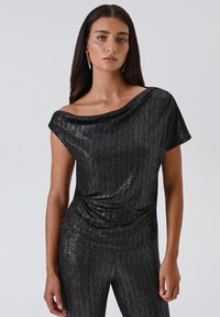 Donna con un top nero lucido off-the-shoulder e pantaloni coordinati, con lunghi capelli scuri e orecchini pendenti dorati, in piedi contro uno sfondo semplice.