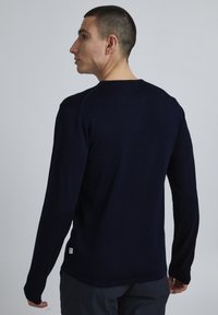 Pulover din material moale, texturat, cu mânecă lungă, de culoare bleumarin, având un guler rotund și un mic eticheta cu logo pe partea din spate, în colțul stâng jos.