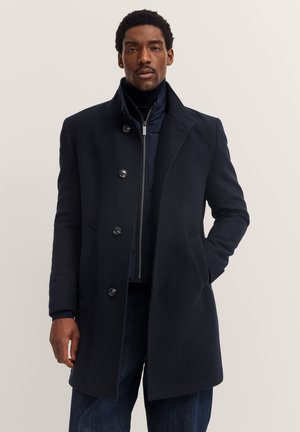 Marineblauer Wollmix-Mantel mit hohem Kragen, fünf Knöpfen, Seitentaschen und strukturiertem Rippdesign, über einer marineblauen Steppjacke getragen.