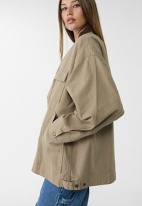 Khaki oversized jakke lavet af bomuld med en ribbet brun krave, to store brystlommer og trykknapsdetaljer.