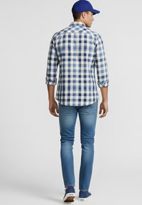 Hombre con camisa a cuadros azul y blanca, vaqueros azules, gorra azul y zapatillas azules de pie de espaldas sobre un fondo liso.