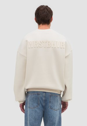 Man draagt een off-white sweatshirt met de tekst "WRSTBHVR" op de rug en blauwe spijkerbroek, staand met de rug naar de camera op een effen achtergrond.