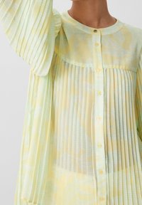 Blusa plissettata gialla chiaro con una texture morbida, dotata di colletto rotondo, chiusura a bottoni e un sottile motivo blu-verde. Design elegante e leggero.