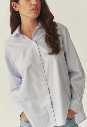 Kobieta o długich brązowych włosach, ubrana w jasnoniebieską koszulę w prążki zapinaną na guziki oraz jasnoniebieskie dżinsy, stoi z rękami w kieszeniach na jednolitym tle.