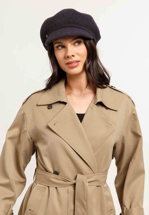 Beige trench coat with a belted waist, double-breasted buttons, and notched collar. Paired with a dark, fitted cap. Smooth texture.