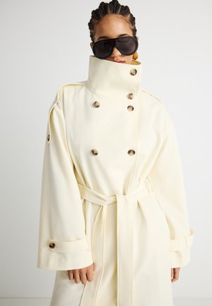 Άτομο που φοράει εκρού διπλό παλτό τύπου trench με ψηλό γιακά, ζώνη στη μέση, μεγάλα κουμπιά και υπερμεγέθια μαύρα γυαλιά ηλίου.