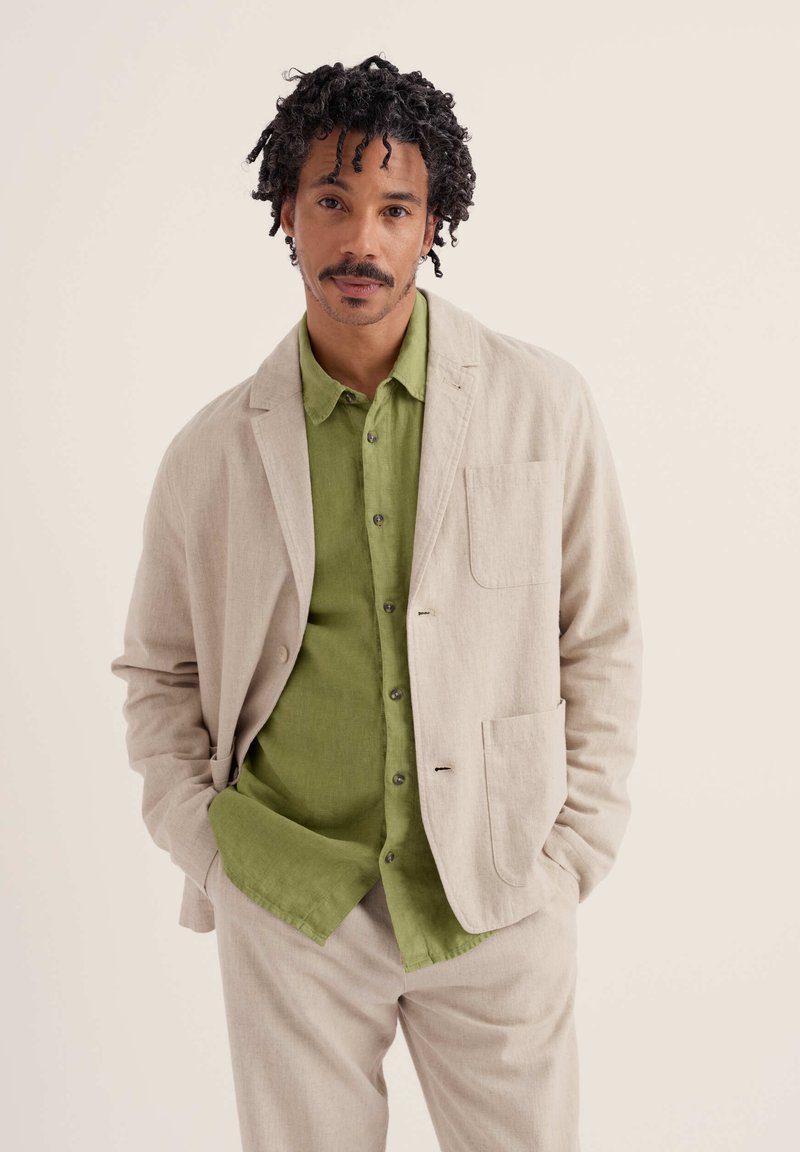Homme aux cheveux bouclés et moustache, portant un costume beige et une chemise verte boutonnée, debout les mains dans les poches devant un fond uni.