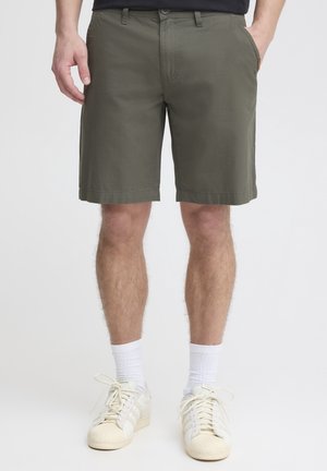 Mand iført olivengrønne shorts til knæet, hvide sokker og off-white sneakers, står med hænderne i lommerne.