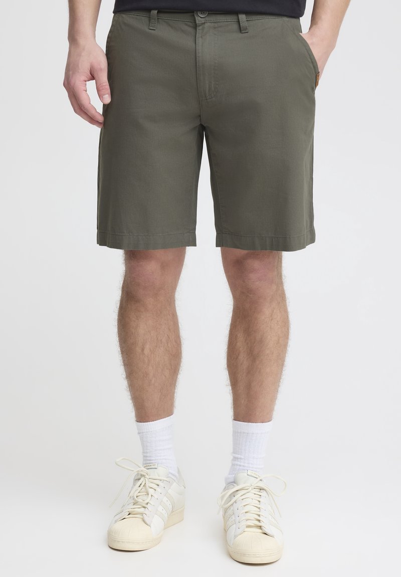 Homme portant un short vert olive jusqu'aux genoux, des chaussettes blanches et des baskets écrues, debout les mains dans les poches.