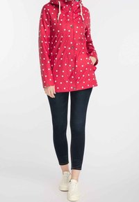 Červená voděodolná bunda s bílými puntíky, zapínání na zip a patentky, nastavitelná kapuce a přední kapsy. V kombinaci s tmavými džíny a bílými teniskami.