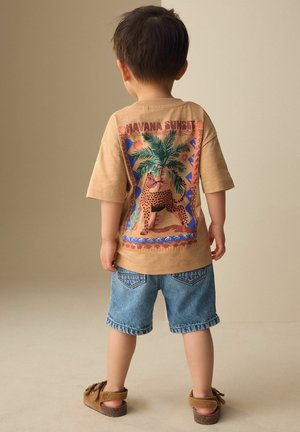 Young child stands facing away wearing tan "Havana Sunset" t-shirt with palm tree and leopard graphic, denim shorts, and brown sandals.